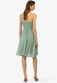 Une robe au genou, à une épaule, de couleur vert clair, avec un corsage froncé et une jupe plissée, dotée d'une texture lisse et d'un minimum de détails.