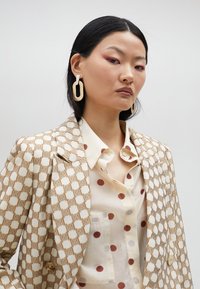 Mönstrad kavaj i krämfärg med en brun geometrisk design, lager på lager över en beige skjorta med maroon prickar. Utstickande örhängen fulländar looken.