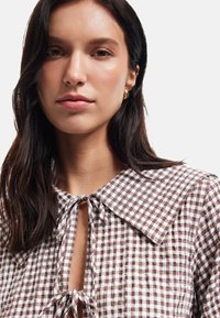 Braun-weiße Gingham-Bluse mit großem Kragen, Vorderbandverschluss und strukturiertem Stoff, der ein kariertes Muster hervorhebt.