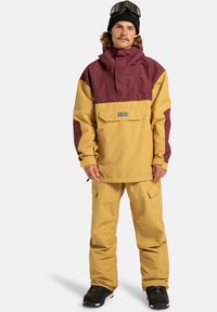 Gelbe und bordeauxrote Ski-Jacke mit einer Fronttasche und passenden gelben Hosen. Eine schwarze Mütze und eine Skibrille vervollständigen das Outfit. Strukturierter Stoff.