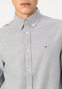 Mees kannab helehallikat nööpidega särki, millel on nööpidega krae ja väikese punase, valge ja sinise ristkülikukujulise logoga rinnal.