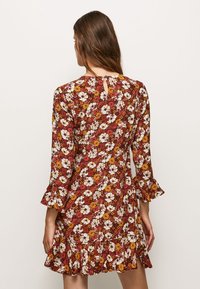 Kvinna som bär en röd blommig klänning med vita och gula blommor, med trekvarts rufsade ärmar och en rufsig fåll, sedd bakifrån.