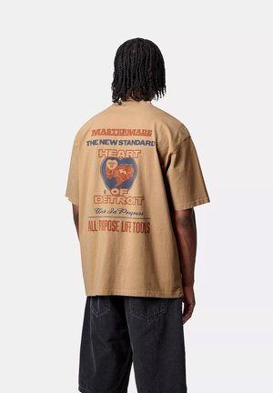Camiseta marrón de gran tamaño con texto gráfico y un emblema circular en la parte posterior. Mangas cortas, cuello redondo, fabricada en algodón.