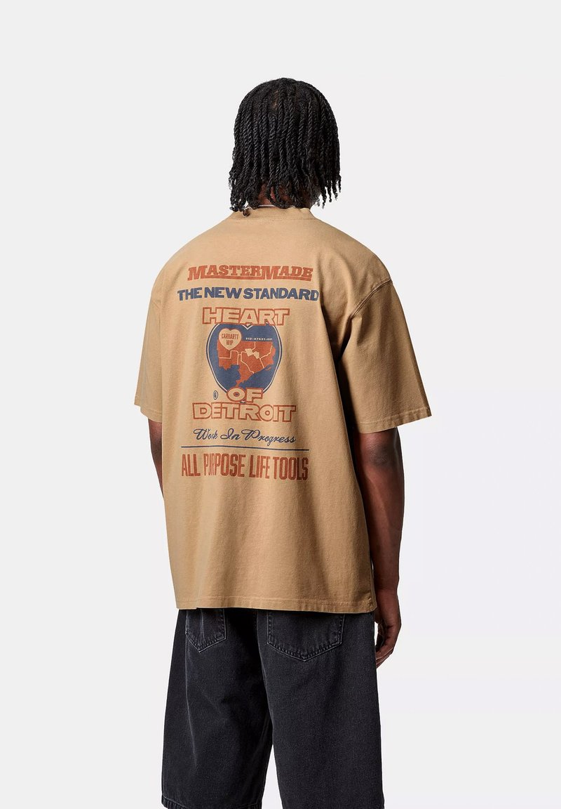 Camiseta marrón de gran tamaño con texto gráfico y un emblema circular en la parte posterior. Mangas cortas, cuello redondo, fabricada en algodón.