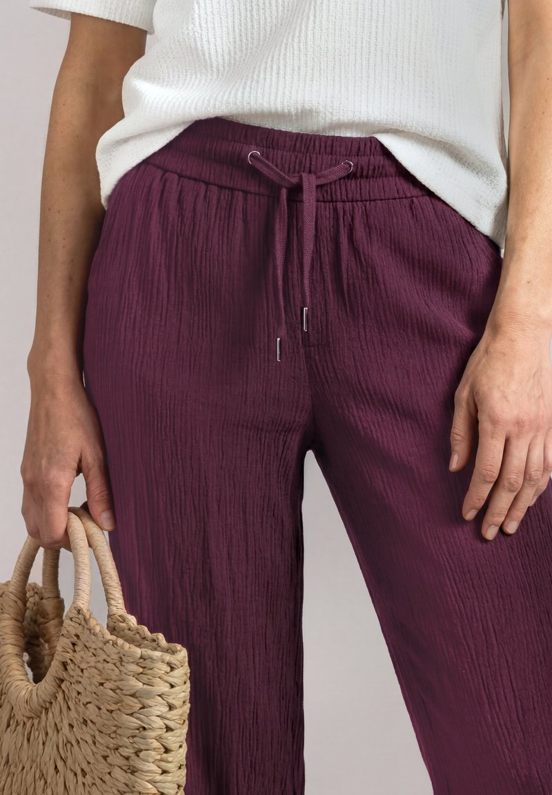 Femme portant un pantalon violet texturé et un haut blanc tenant un sac à main en paille tissée avec des poignées rondes.