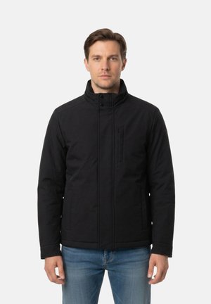 Uomo con giacca nera con zip e colletto a listino e jeans blu, in piedi davanti a uno sfondo chiaro e uniforme.