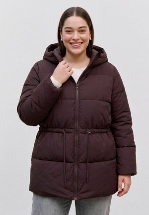 Femme souriante portant une doudoune marron foncé à capuche avec zip à l'avant et cordon à la taille, debout devant un fond clair uni.