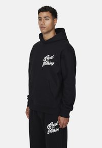 Czarna bluza z kapturem, z przednią kieszenią kangurką, z białym napisem "Good for Nothing" na klatce piersiowej oraz dopasowane czarne spodnie z podobnym napisem.