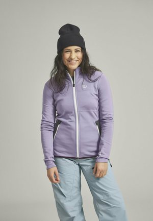 Purple softshell jacket with white zipper and side pockets, paired with light blue pants. Black knit beanie completes the look.