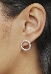 Pendiente de aro de plata con una superficie lisa y brillante, de diseño circular sencillo y un pequeño cierre de gancho, llevado en una oreja.