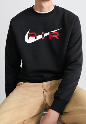 Person trägt schwarzen Pullover mit weißem Nike-Swoosh und rotem "AIR"-Schriftzug, kombiniert mit beigen Hosen, sitzt vor einfarbigem Hintergrund.