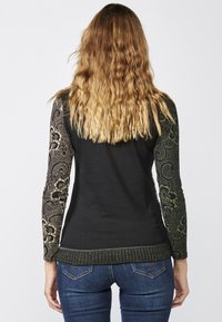 Schwarzes Langarmoberteil mit gemusterten grünen und beigen Spitzeärmeln, geripptem Saum und tailliertem Design, kombiniert mit blauen Jeans.