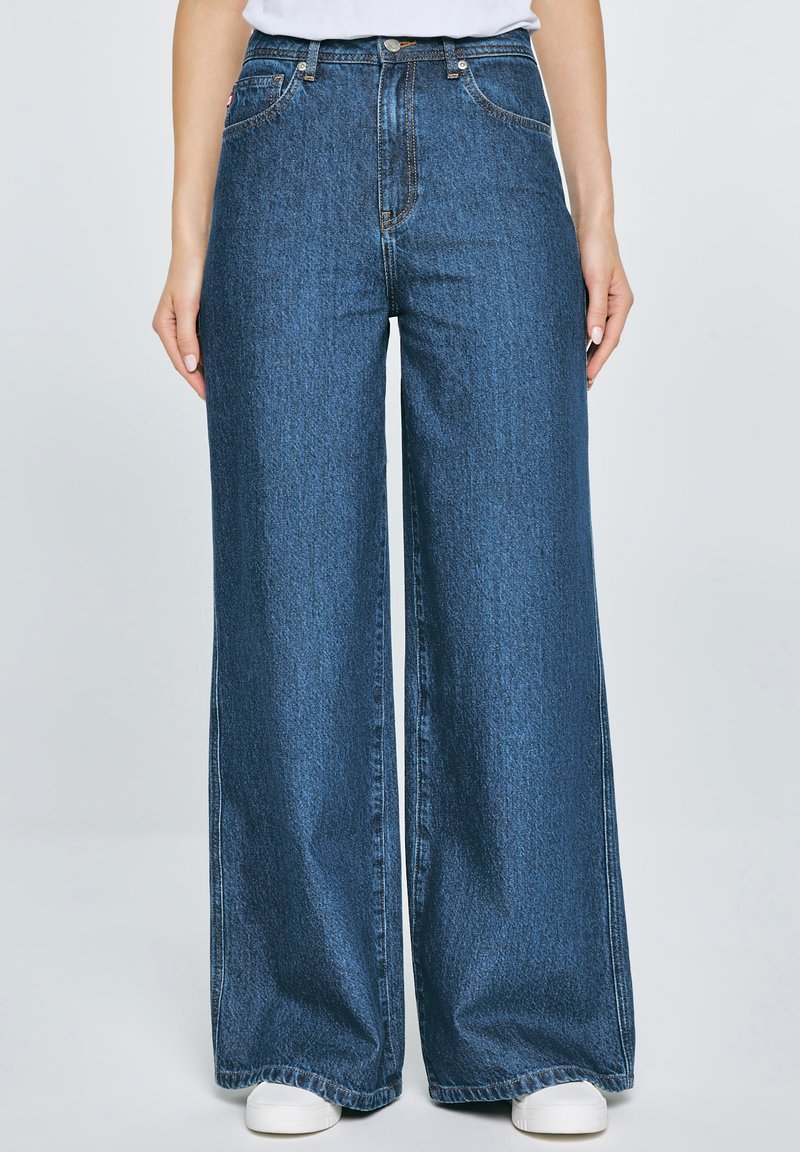 Wide-leg blue denim jeans with a high waist, featuring five pockets and contrasting stitching, paired with white sneakers.