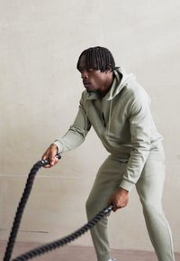 Athletisk person i lys grøn hættetrøje og matchende sweatpants griber sorte battle ropes, fokuseret på træningen mod en neutral baggrund.