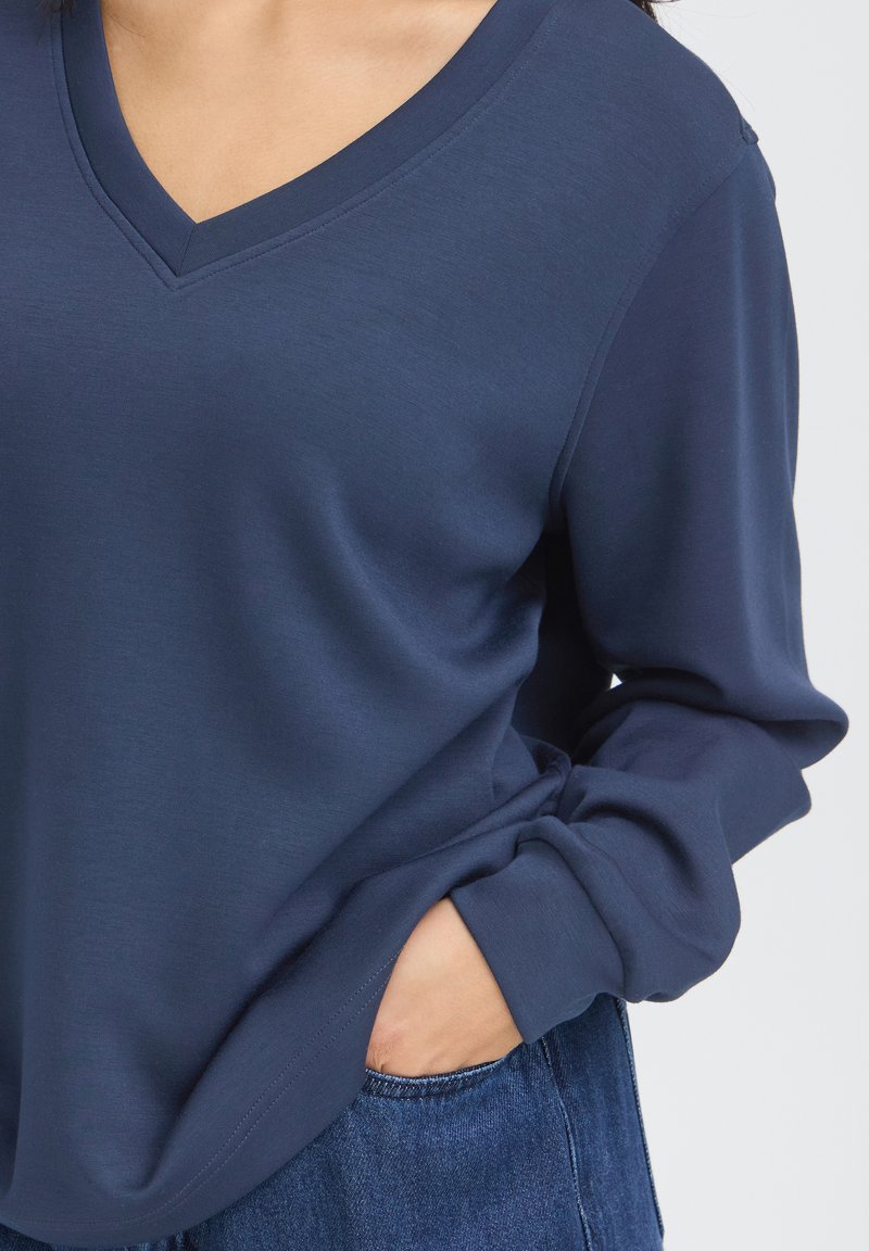 Personne portant un tee-shirt bleu marine à manches longues avec un col en V, une main dans la poche d'un jean bleu.