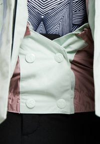 Jasnozielona kurtka o dwurzędowym kroju, z pięcioma dużymi białymi guzikami; wewnątrz różowa podszewka oraz czarno-biały geometryczny wzór.