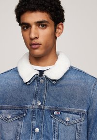 Giubbotto di jeans con colletto in pelliccia sintetica bianca, dotato di due tasche sul petto, bottoni argentati e una piccola etichetta con logo sul davanti.