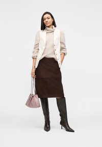Femme portant un pull à col roulé beige, un gilet crème, une jupe marron foncé, des bottes noires montantes, tenant un sac à cordon mauve, debout devant un fond blanc.