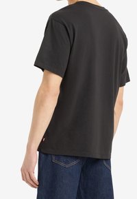 Man klädd i en enkel svart kortärmad T-shirt och mörkblå jeans, sedd bakifrån mot en vit bakgrund.