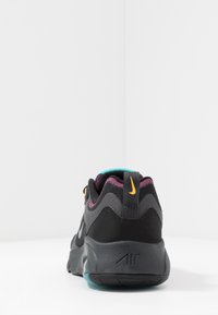 Takana näkökulma mustasta Nike Air -lenkkarista, jossa on violetit, teal-vihreät korostukset ja keltainen logo kantapäässä, valkoista taustaa vasten.