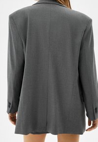 Frau, die einen übergroßen grauen Blazer trägt, von hinten gesehen, mit nackten Beinen, die unter dem Jackenbund hervorschauen.