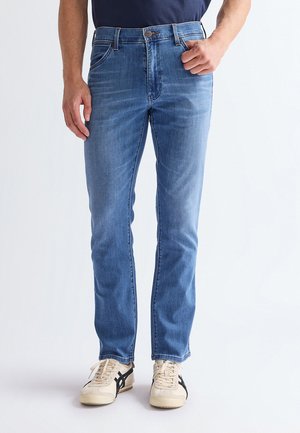 Man draagt medium blauwe slim-fit jeans en crème sneakers met zwarte strepen, staand met één hand bij de zak.