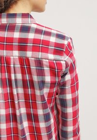 Koszula flanelowa w kratę w kolorach czerwonym, białym i granatowym, wyposażona w tylny kawałek materiału, kołnierzyk zapinany na guziki oraz długie rękawy z mankietami.