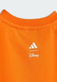 Oranžové bavlnené tričko s golierom a šitými okrajmi. Zdobené bielym logom Adidas a textom Disney nižšie, hladká textúra a krátke rukávy.