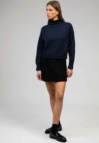 Pull en maille bleu marine foncé avec col roulé et ourlet côtelé, associé à une mini-jupe noire texturée et des mocassins en cuir verni noirs avec des chaussettes courtes.