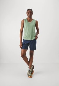 Woman standing with legs crossed, wearing green sleeveless top, navy shorts, gray socks, and green hiking shoes on plain background.