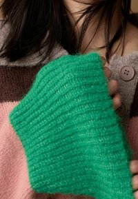 Gros plan d'une personne portant un pull en laine rayé tenant la manche verte d'un autre vêtement tricoté contre sa poitrine.