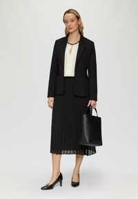 Schwarze Blazer mit Revers über einer cremefarbenen Bluse mit einem Schlüsselloch-Ausschnitt, kombiniert mit einem plissierten schwarzen Rock und schwarzen spitzen Schuhen.