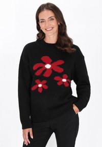 Schwarzer Pullover mit roten Blumenmustern und weißen Zentren, mit ribbeligem Halsausschnitt und Bündchen. Modell posiert mit Händen in den Taschen.