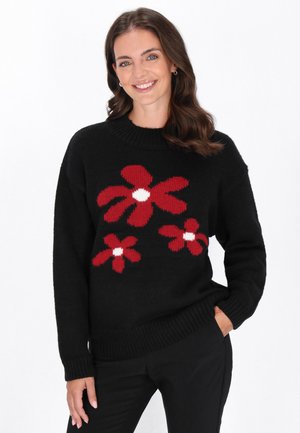 Schwarzer Pullover mit roten Blumenmustern und weißen Zentren, mit ribbeligem Halsausschnitt und Bündchen. Modell posiert mit Händen in den Taschen.