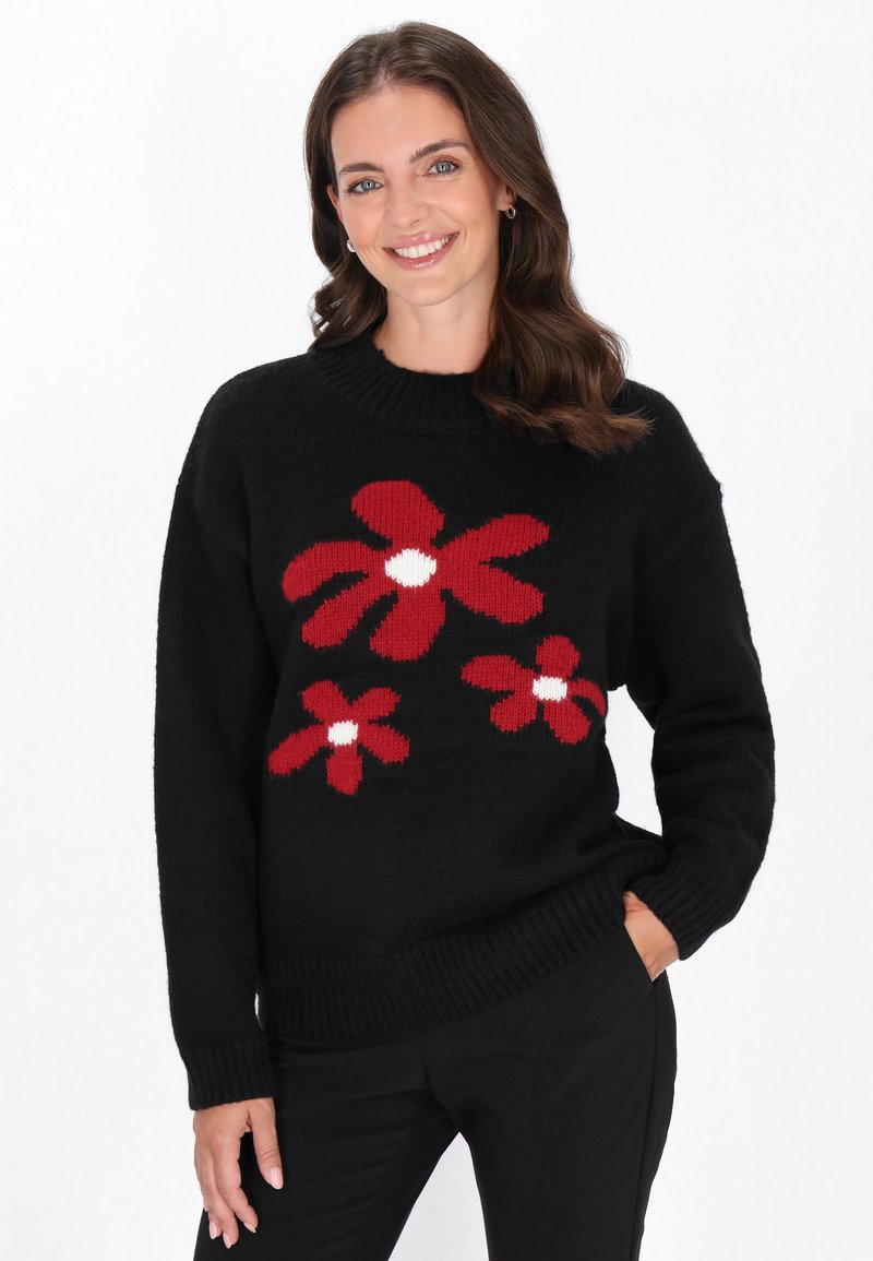 Schwarzer Pullover mit roten Blumenmustern und weißen Zentren, mit ribbeligem Halsausschnitt und Bündchen. Modell posiert mit Händen in den Taschen.