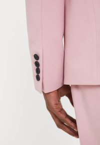 Manche d'un costume rose ajusté, avec quatre boutons noirs et un tissu légèrement texturé, montrant un gros plan d'une main reposant contre la manche.