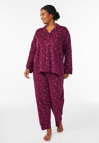 Tummanpunainen pyjama-asu, jossa valkoiset sydänkuviot, nappipaita kauluksella ja rentoistukkaiset housut. Materiaali puuvillaa.