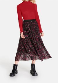 Haut rouge à manches longues associé à une jupe plissée noire à motif géométrique rose. Les chaussures comprennent des bottines noires à lacets.