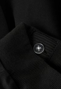 Sort stof med en ribbet struktur, prydet med en klar, firehuls knap og en glat finish. Knapen er syet fast til stoffet.