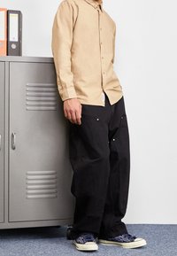 Camisa de botones de manga larga color beige claro, pantalones cargo negros holgados con bolsillos y zapatillas con patrón en color marino. El fondo presenta un casillero gris.