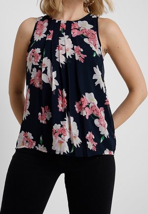 Mouwloze marineblauwe blouse met roze en witte bloemenprint, geplooide voorkant, gedragen met zwarte broek.