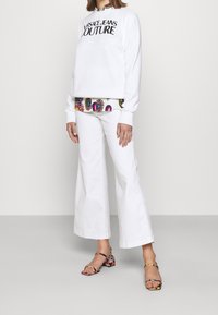 Bílá mikina s nápisem "Versace Jeans Couture" v kombinaci s bílými zvonovými džíny. Sandály mají vícebarevný květinový vzor na chunky podpatku.