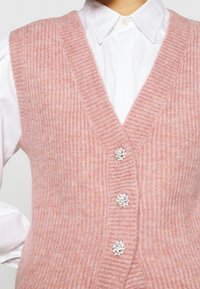 Gilet a coste rosa con scollo a V profondo, con tre bottoni floreali decorativi sul davanti. Indossato sopra una camicia bianca con colletto.