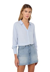 Blusa azzurro chiaro, con bottoni e maniche lunghe, colletto e texture morbida, abbinata a una minigonna di jeans chiari.