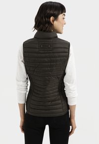 Donkergroene geïsoleerde vest met ribbeltextuur, hoge kraag en drukknopaccenten. Draagt over een witte lange mouw shirt.