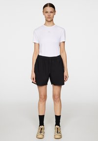 Vit kortärmad t-shirt med en logotyp, parat med svarta shorts med dragsko och beigea träningsskor. Enkelt, funktionellt design med en avslappnad passform.