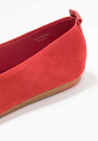Chaussure plate en suede rouge avec un bout arrondi, un petit talon et une accent latéral texturé. L'intérieur a une doublure rouge lisse et une étiquette de taille.