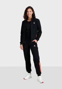 Tuta sportiva nera con accenti arancioni, composta da giacca con cappuccio con zip e pantaloni con vita elastica. Scarpe da ginnastica bianche.