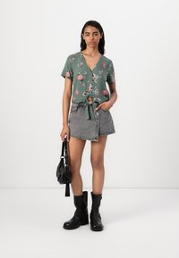 Blusa floreale verde con maniche corte e dettagli annodati davanti, abbinata a una gonna di jeans grigia. Stivaletti neri alla caviglia e una piccola borsa nera.