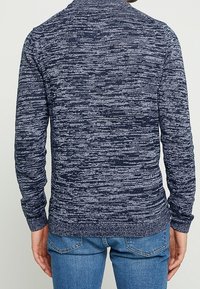 Un pull tricoté bleu marine avec un motif texturé et marbré. Il présente un col rond et des poignets côtelés. Porté avec un jean bleu clair.